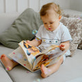 A baby sitting on a light gray sofa, reading a colorful picture book. The baby is wearing a white short-sleeve romper with small hands-mocked details.