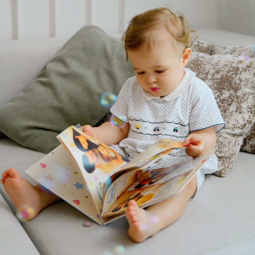 A baby sitting on a light gray sofa, reading a colorful picture book. The baby is wearing a white short-sleeve romper with small hands-mocked details.